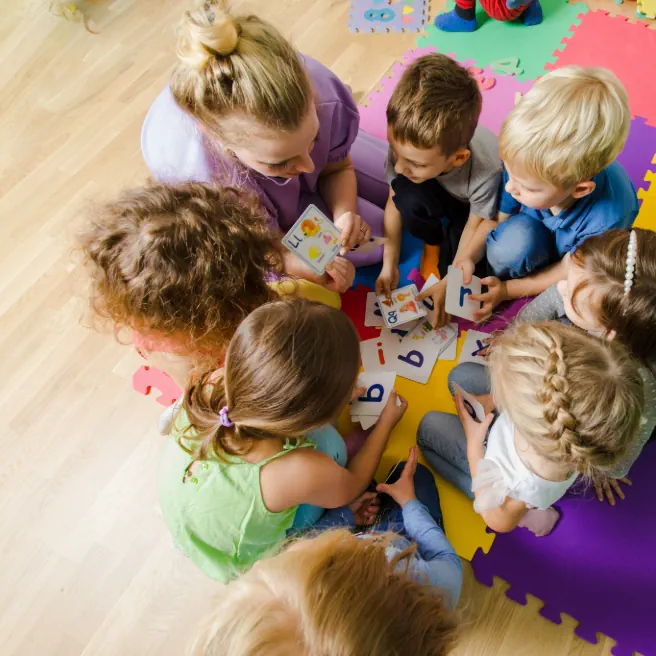 Éducatrice entourée d’enfants en crèche, jouant et apprenant avec des cartes éducatives – illustration du débat sur l’éducation positive et la petite enfance.