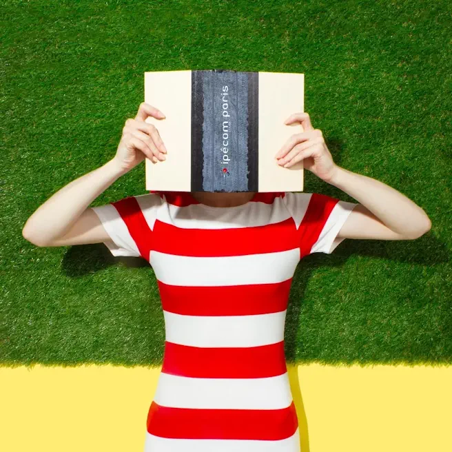 Élève en T-shirt rayé tenant un livre Ipécom Paris devant son visage, allongée sur un fond vert.